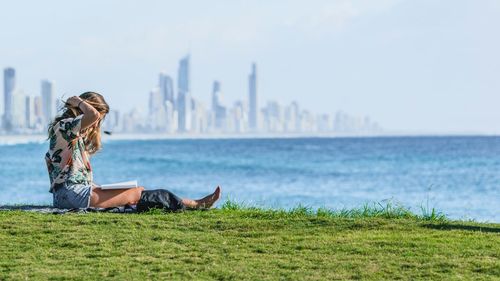 Side view of woman reading book while sitting on grass by sea in city