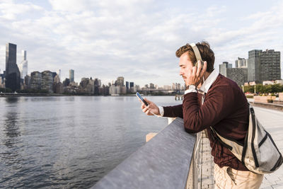 Young businessman wearing headphones and listening to music by hudson river in new york city, usa