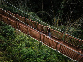 High angle portrait of woman standing on footbridge over trees in forest