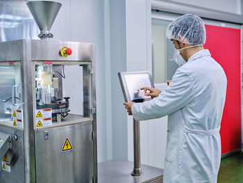Side view of male chemist in uniform operating capsule filling machine at pharmaceutical manufacturing plant