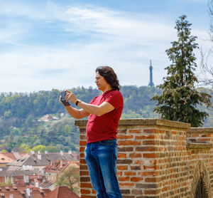 A man stands on a brick wall and photographs the panorama of prague. person