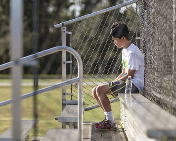 Thoughtful teenage boy sitting on bench against chainlink fence at playing field