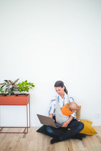 Full length of working mother using laptop while sitting with daughter against wall at home office