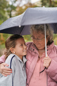 Grandmother embracing granddaughter while sharing umbrella with her in park during rainy season