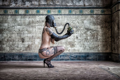 Full length of sensuous naked woman wearing gas mask against wall