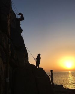 Silhouette people climbing rock against sky during sunset