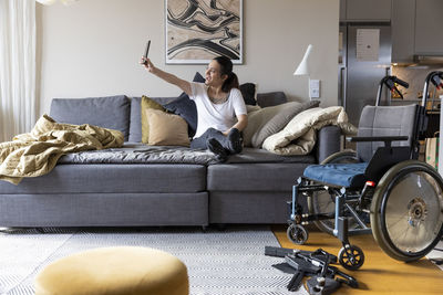 Happy woman with paraplegia taking selfie through smart phone while sitting on sofa in living room at home