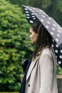 Portrait of a young woman with dotted umbrella standing against trees