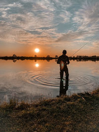 Rear view of man standing by lake against sky during sunset