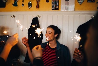 Smiling young woman holding burning sparkler while enjoying with multi-ethnic friends in restaurant during dinner party