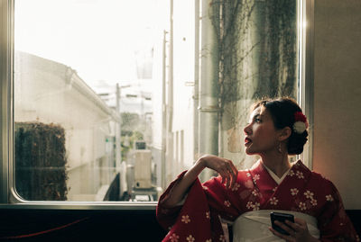 Woman looking through window while using mobile phone on sofa at home