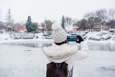 Back view of backpacker woman taking picture with mobile phone, standing in front of frozen lake