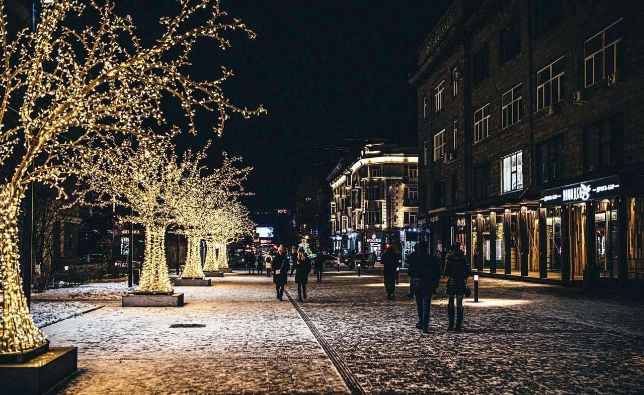 PEOPLE ON ILLUMINATED STREET BY BUILDINGS AT NIGHT