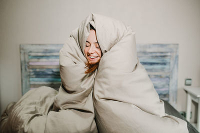 Smiling woman wearing blanket sitting on bed at home