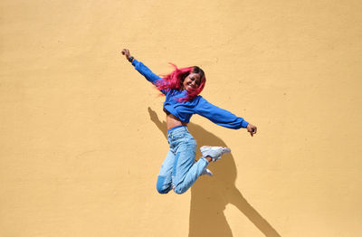 Full body of overjoyed young african american hipster female with pink hair wearing blue shirt and jeans jumping high above ground against yellow wall