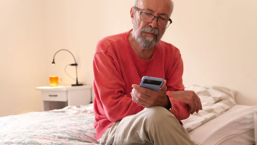 Senior man using mobile phone while sitting on bed at home