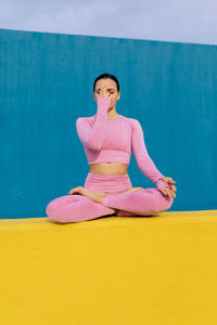 Peaceful female in pink activewear sitting in padmasana with namaste hands and meditating during yoga session on blue and yellow background