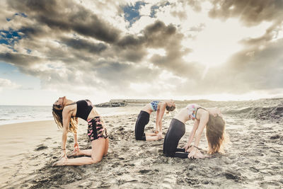 Women doing stretching at beach against sky suring sunset