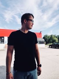 Young man looking away while standing on street against sky