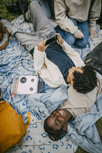 High angle view of teenage boy using smart phone while lying on male friend in public park