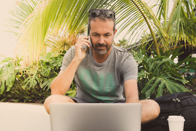 Mature man talking over phone while sitting against plants with laptop