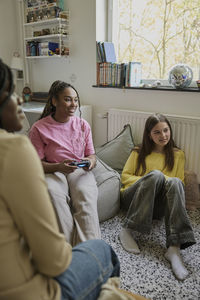 Teenage girl playing video game by female friends sitting at home during weekend