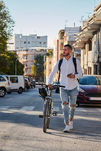 Full body of serious adult bearded male in trendy ripped jeans with backpack checking information on mobile phone while walking with bicycle on paved urban street