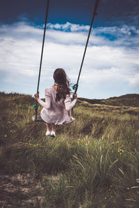 Rear view of woman on swing over grassy field against cloudy sky
