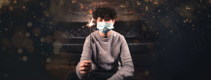 Portrait of man wearing mask holding sparkler sitting at home