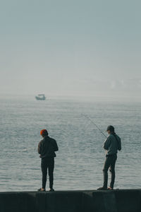 Rear view of men standing by sea against sky while fishing