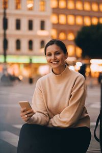 Portrait of smiling young woman using phone while sitting in city