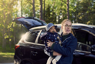 Young woman carrying baby boy while standing against car in forest