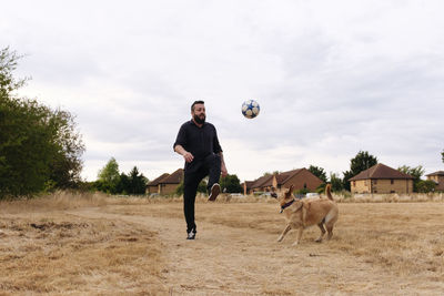 Man playing with dog kicking soccer ball in field at sunset