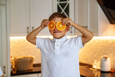 A funny boy holding fresh orange slice on eyes in kitchen