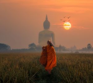 Rear view of monk walking towards buddha statue on field against sky during sunset