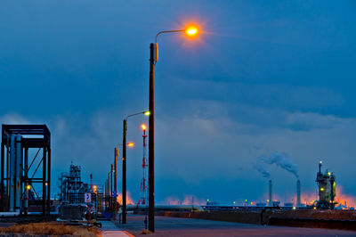 Illuminated street by factory against sky at dusk