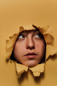 Pensive youthful female teen with watching out from ripped hole in yellow paper in studio