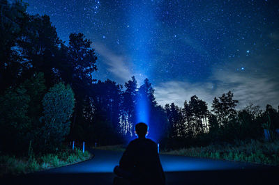 Rear view of person holding flashlight sitting against trees at night