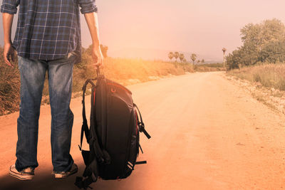 Low section of man with backpack standing on dirt road against sky