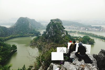 Rear view of people painting on canvas against river and mountains during foggy weather