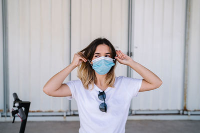 Serious young female student in casual outfit and medical mask looking at camera while standing near electric scooter against metal wall