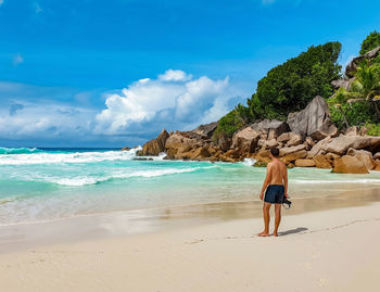 Rear view of shirtless young man standing on tropical sandy beach, holding camera