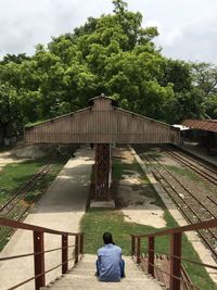 Rear view of man sitting on railroad station against trees