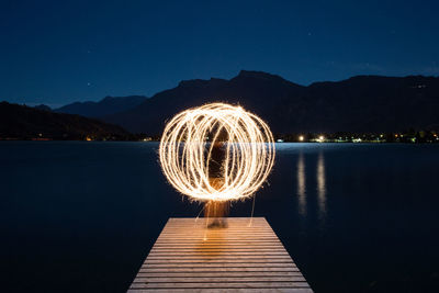 Person spinning illuminated wire wool while standing on pier against lake at night