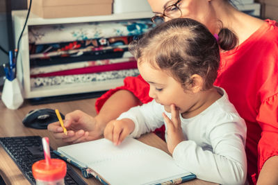 Mother teaching cute girl to write in book while sitting on table at home