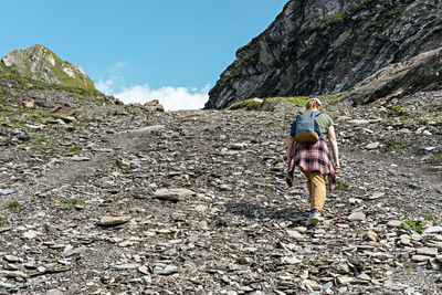 Rear view young woman with backpac walking uphill along rocky trail in mountain hike, hiking 