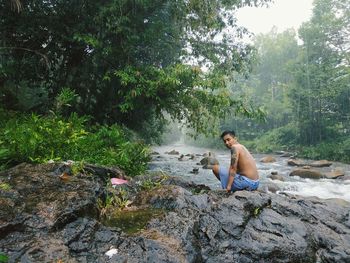 Rear view of shirtless man sitting on rock in forest by stream