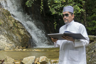 Mature man reading book against waterfall in forest