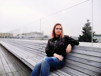 Young woman sitting on bench against sky