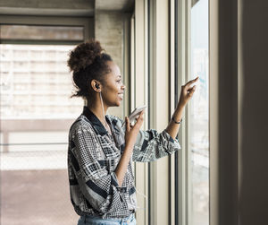 Young businesswoman with smart phone looking out window in office